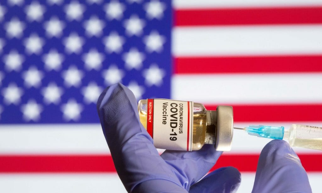 A woman holds a small bottle labeled with a "Coronavirus COVID-19 Vaccine" sticker and a medical syringe in front of displayed USA flag in this illustration taken, October 30, 2020. REUTERS/Dado Ruvic