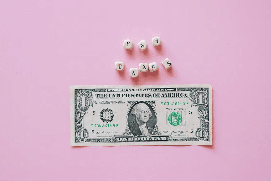 Wooden letter cubes spelling “PAY TAXES” above a U.S. one-dollar bill on a pink background.