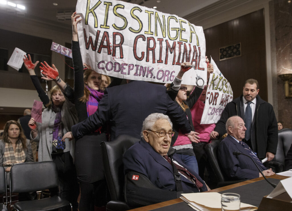 Henry Kissinger sitting down with "Kissinger War Criminal" banner behind him
