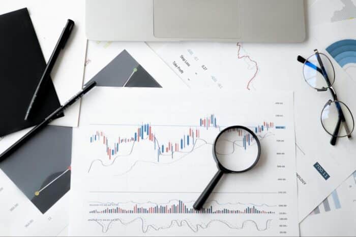 Desk setup with a laptop, trading charts, graphs, a clock, and financial documents, symbolizing active trading and market analysis.