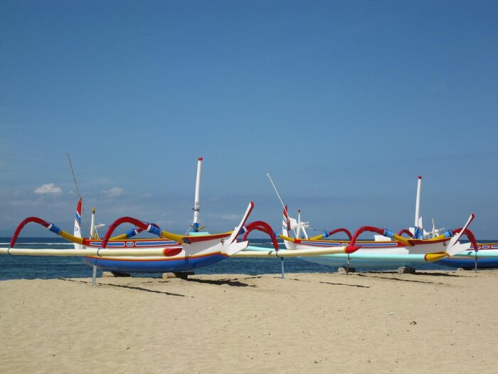 Traditional fishing boats with colorful sails lined up on the sandy shore of Sanur Beach, Bali, under a clear blue sky.