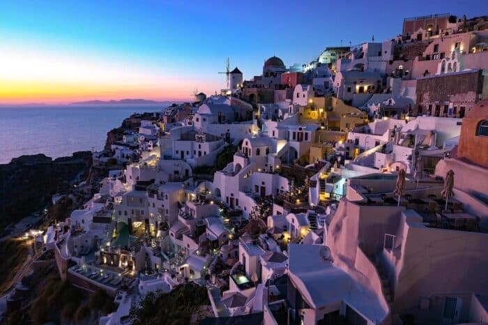 Stunning sunset view over Santorini, Greece, with iconic whitewashed buildings, glowing lights, and the Aegean Sea in the background.