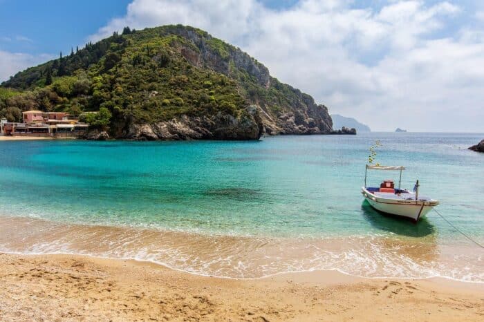 A beautiful beach in Corfu, Greece, with turquoise waters, sandy shore, and a small boat anchored near lush green hills under a clear sky.