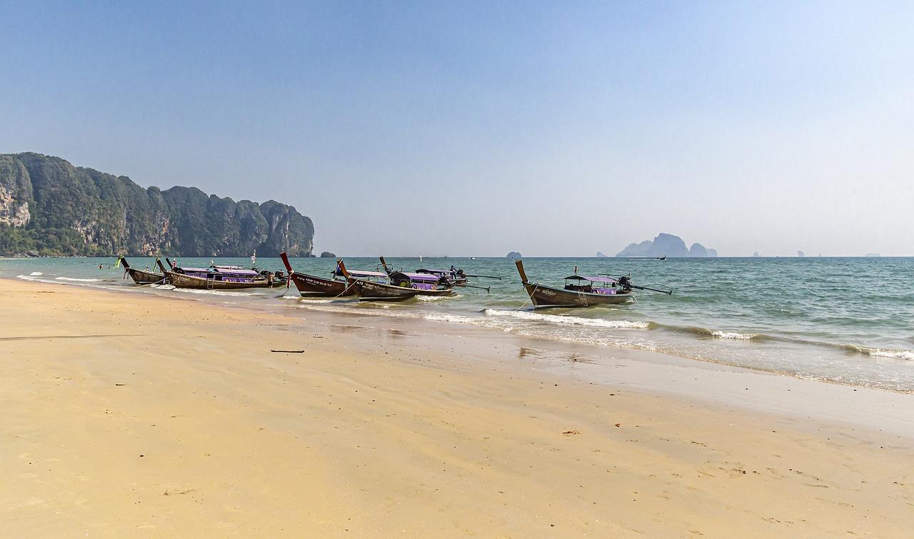 Long-tail boats anchored on the sandy shore of Ao Nang Beach, Krabi, Thailand, with limestone cliffs and calm blue waters in the background.