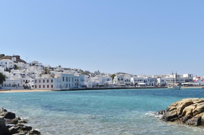 Scenic view of Mykonos, Greece, featuring white buildings along the coastline, crystal-clear blue waters, and rocky shoreline under a bright blue sky.