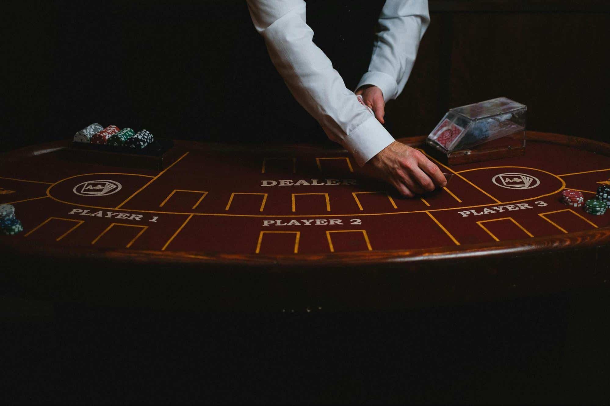 A casino dealer in a white shirt and black vest dealing cards at a blackjack table, with poker chips and playing cards spread across the table.