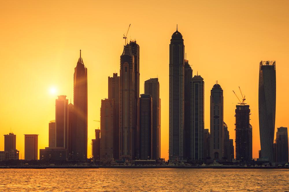 A dramatic sunset over the Dubai Marina skyline, featuring tall modern skyscrapers silhouetted against an orange sky, reflecting off the water in the foreground.