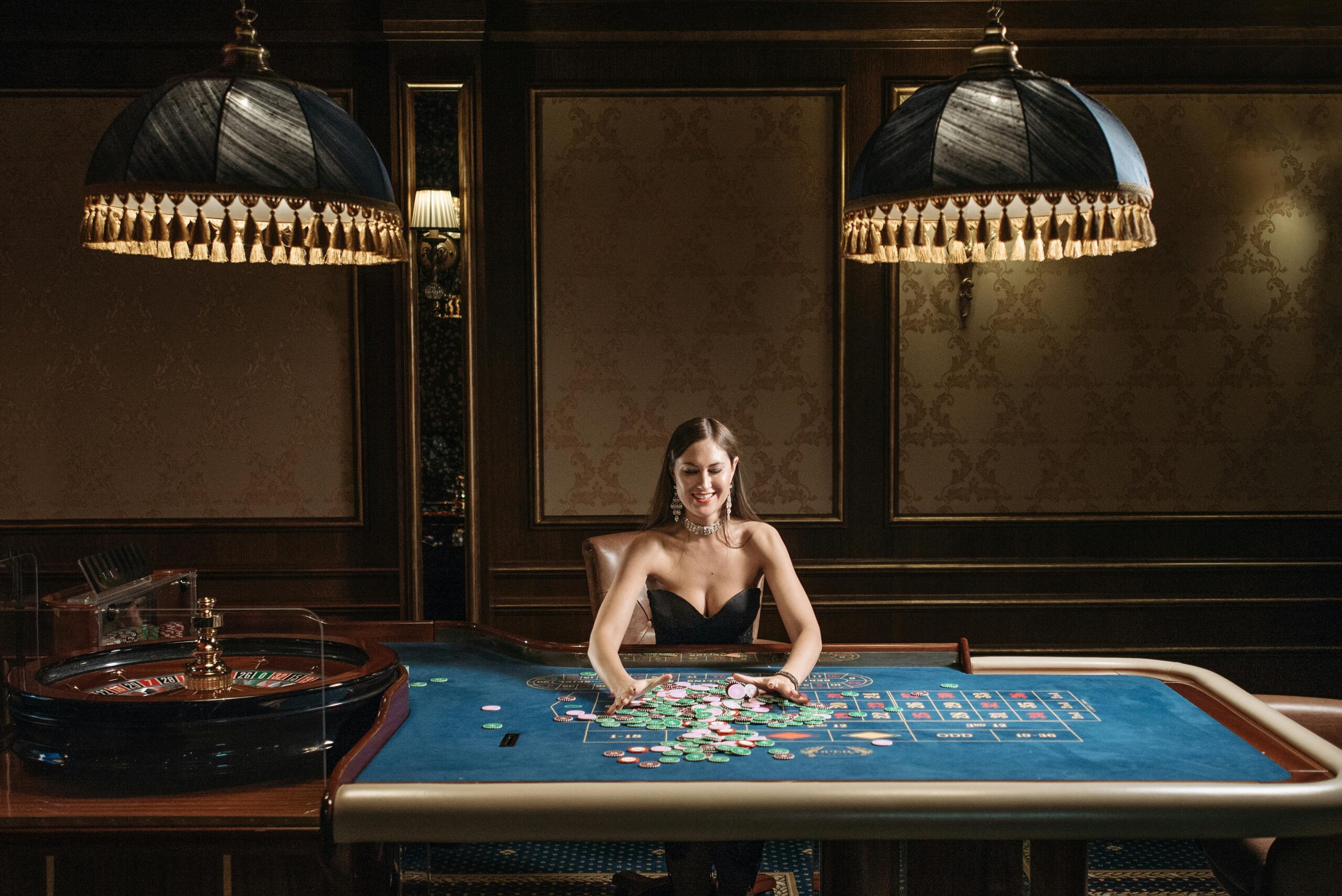 A female casino dealer sits behind a roulette and card table inside a dimly lit Macau casino.
