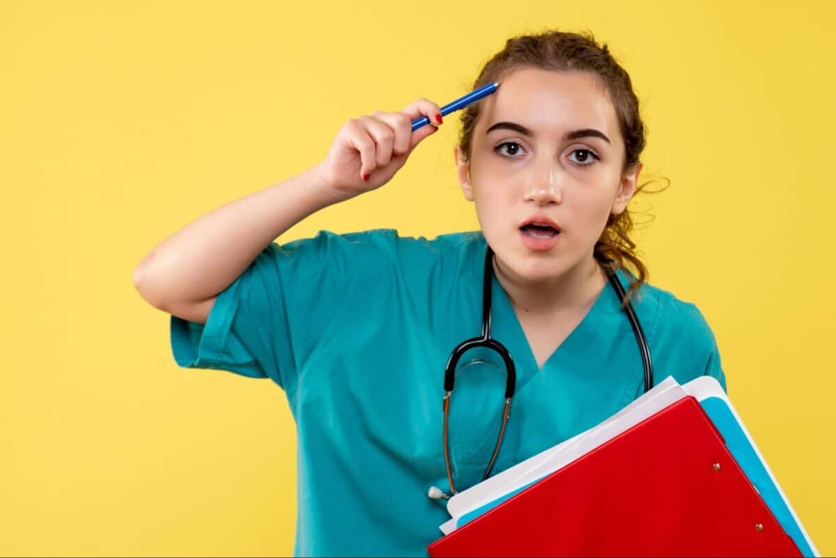 A medical student in scrubs holding folders and a pen, appearing thoughtful against a yellow background.