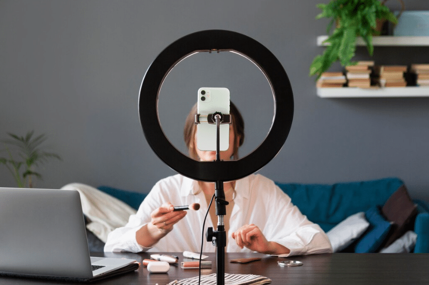 A content creator sitting at a desk with a ring light and smartphone setup, holding a makeup brush while preparing to film a video.