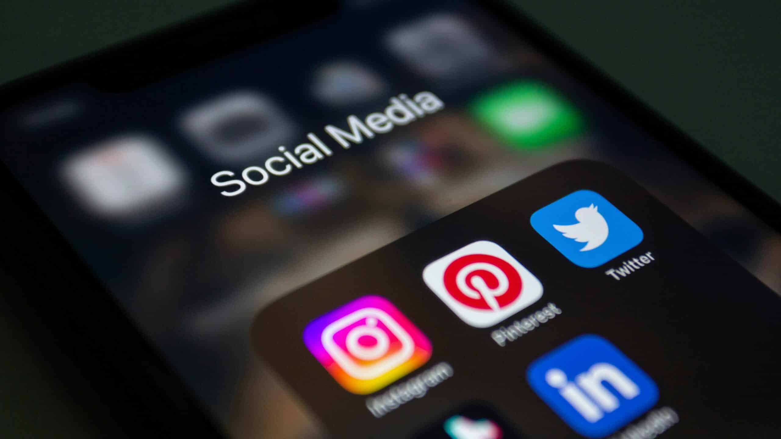 A close-up, angled shot of a smartphone screen displaying a folder labeled "Social Media," which contains apps for Instagram, Pinterest, Twitter, and LinkedIn. The background is dark and slightly blurred, emphasizing the vibrant icons on the display.