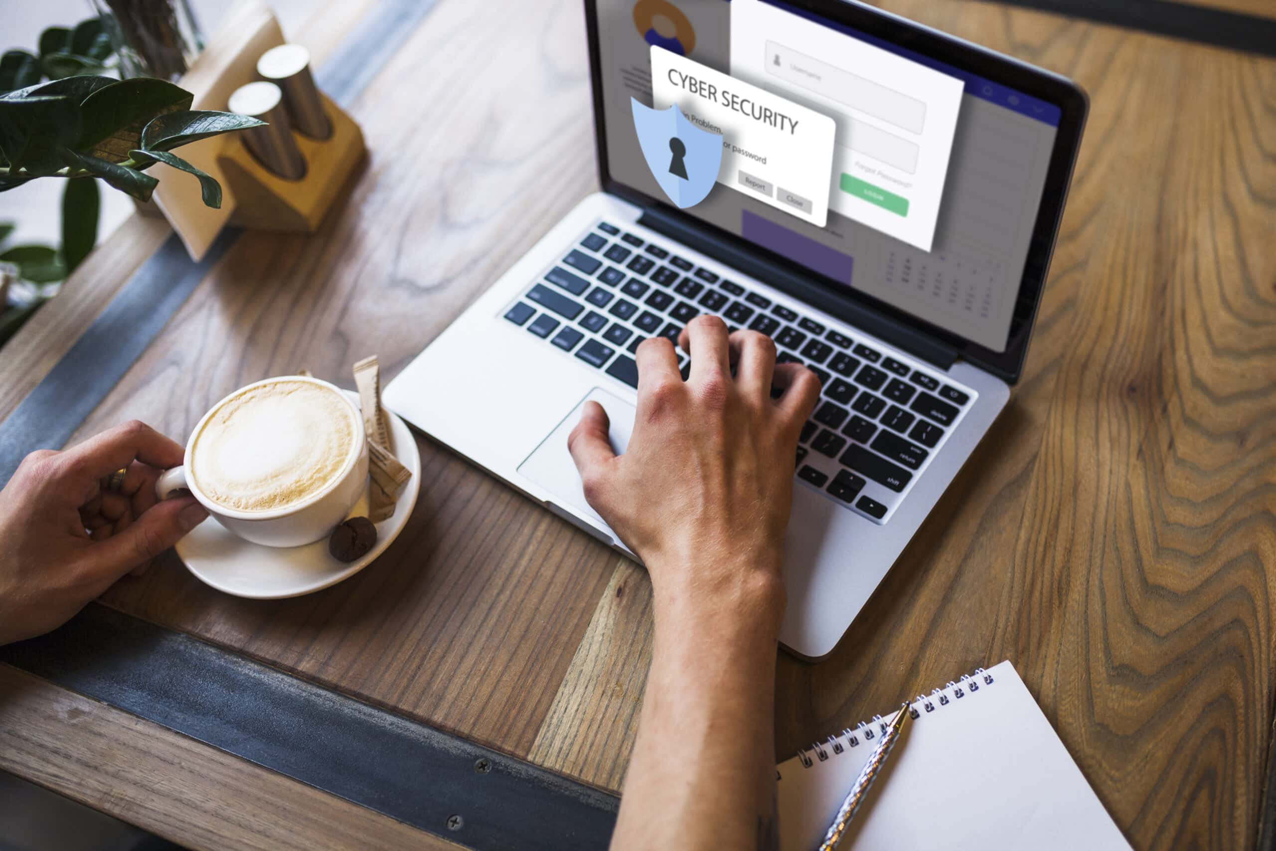 A person typing on a laptop at a wooden desk with a latte nearby, while the laptop screen displays a cyber security interface featuring a padlock icon and login form.
