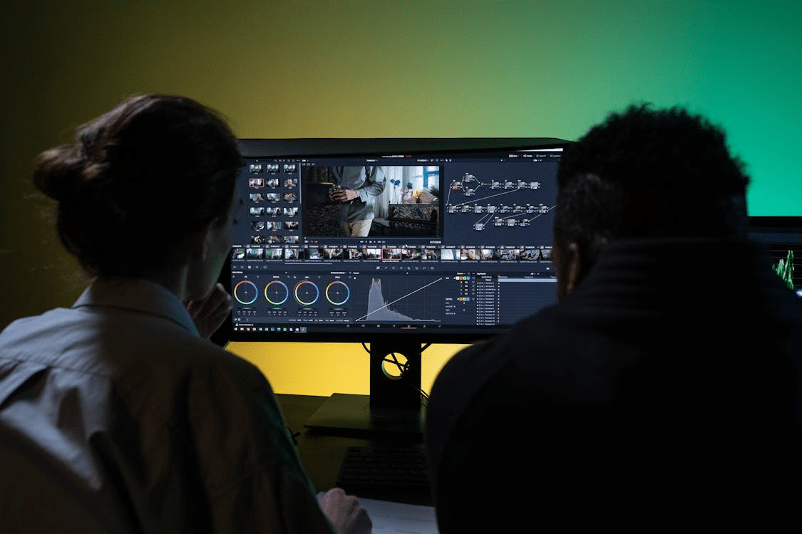 Two video editors sitting in front of a large computer monitor, reviewing a video project timeline, node-based editing workflow, and color grading wheels in a dark studio with green and yellow ambient lighting.