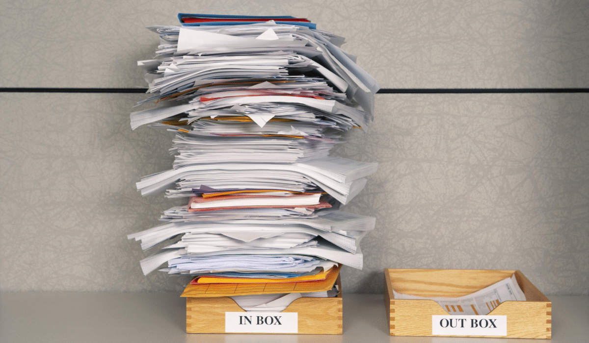 A tall, disorganized stack of papers overflowing from an 'IN BOX' on an office desk, contrasting with an empty 'OUT BOX'